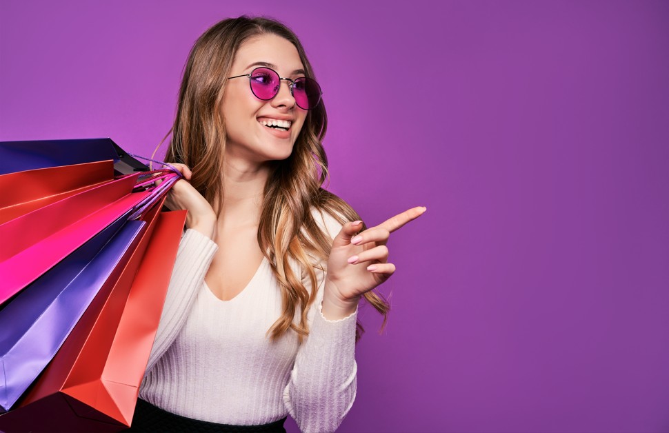 Une jeune femme souriante tenant des sacs de shopping à l'épaule et pointant un doigt sur le côté