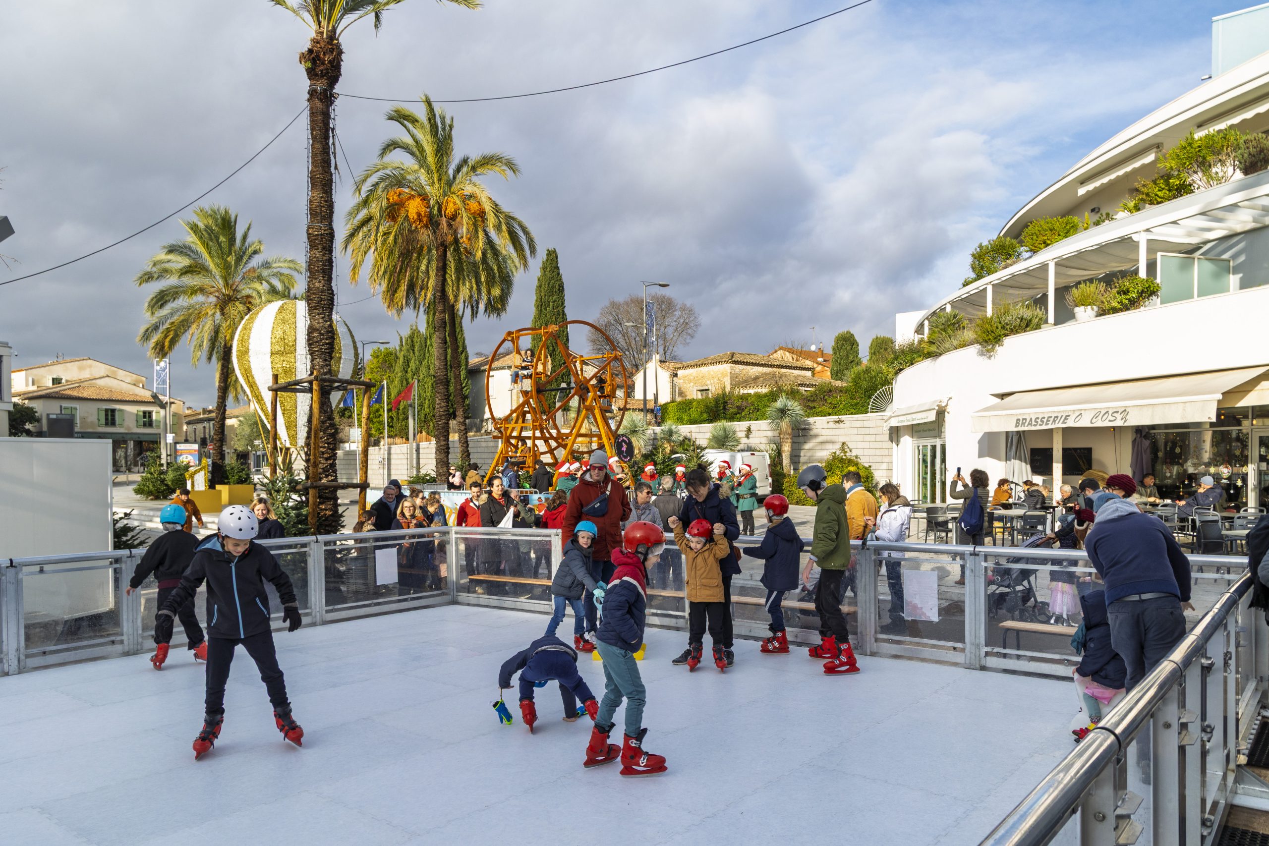 Des enfants sur la patinoire au premier plan, une grande roue en bois et une immense montgolfière en arrière-plan