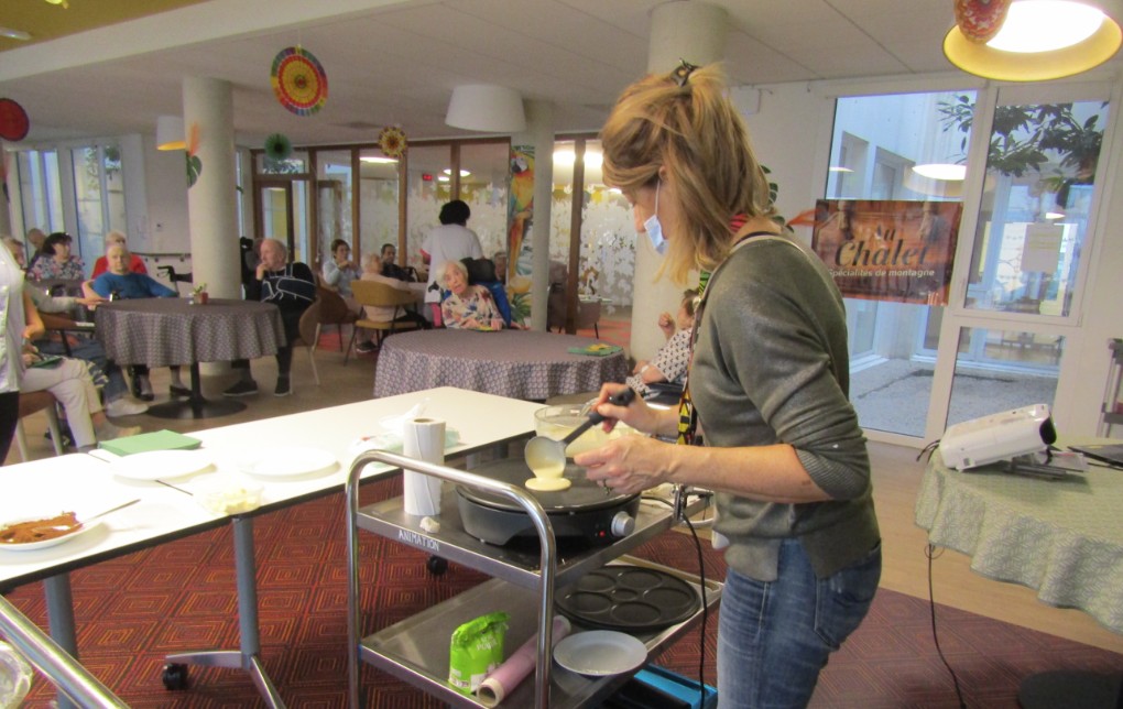 Une femme vue de dos en train de faire des crêpes, avec en arrière-plan des résidents d'un EHPAD de Castelnau-le-Lez
